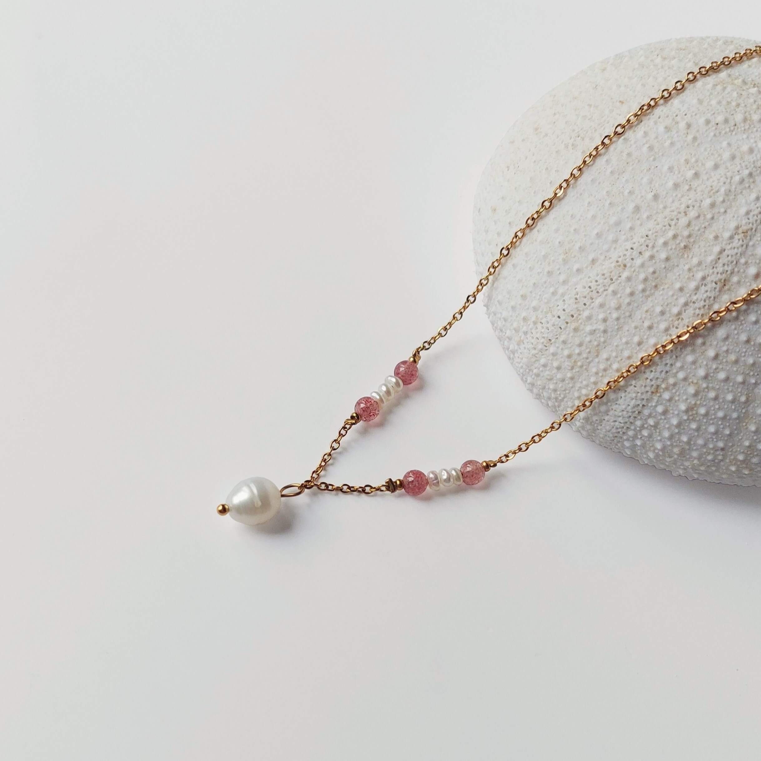 Collier Euphyllia en quartz fraise et petites perles d'eau douce sur fine chaîne dorée en acier inoxydable, avec une perle d'eau douce baroque en pendentif, posé contre un oursin blanc sur fond gris clair.