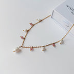 Collier Meandrina quartz fraise en acier inoxydable doré avec breloques de perles d'eau douce baroques et pierres de quartz fraise roses alternées, posé sur fond blanc avec une boîte à bijoux Iridescence Bijoux en arrière-plan.