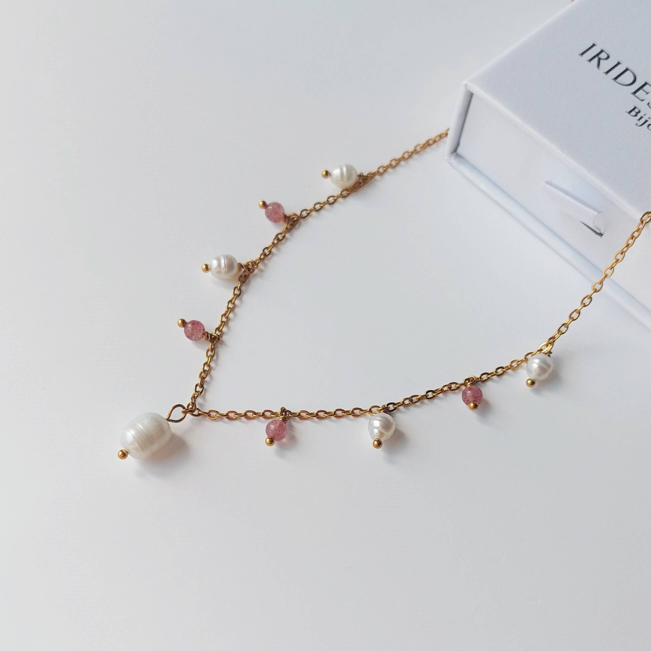 Collier Meandrina quartz fraise en acier inoxydable doré avec breloques de perles d'eau douce baroques et pierres de quartz fraise roses alternées, posé sur fond blanc avec une boîte à bijoux Iridescence Bijoux en arrière-plan.