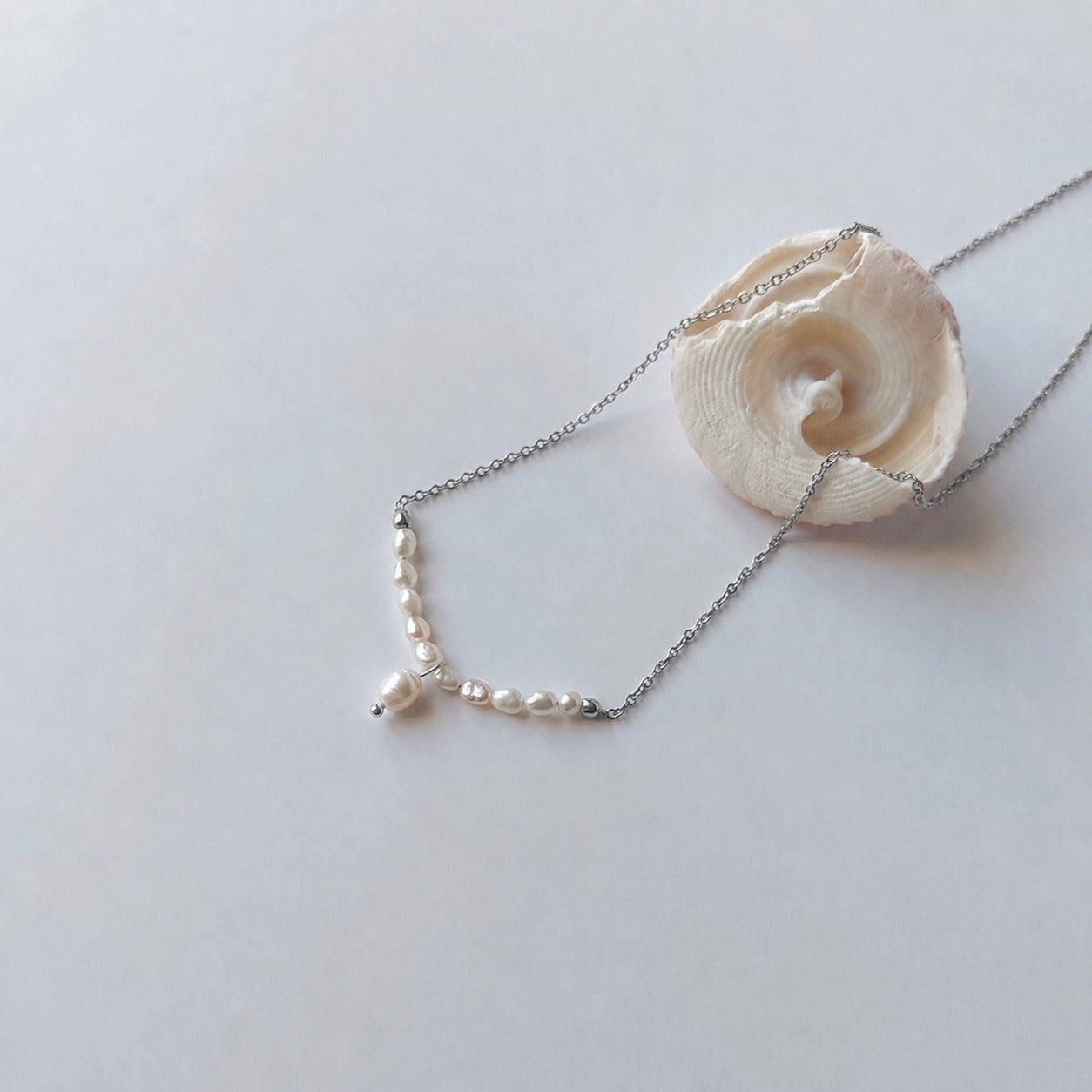 Collier fin sur chaîne argentée composé d'une rangée de petites perles d'eau douce blanches baroques en arc, avec une perle plus grande en pendentif central, posé sur fond gris clair aux côtés d'un coquillage en spirale blanc.