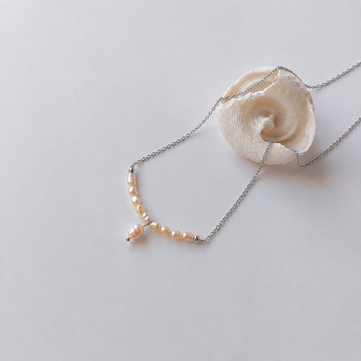 Collier fin sur chaîne argentée composé d'une rangée de petites perles d'eau douce oranges baroques en arc, avec une perle plus grande en pendentif central, posé sur fond gris clair aux côtés d'un coquillage en spirale blanc.
