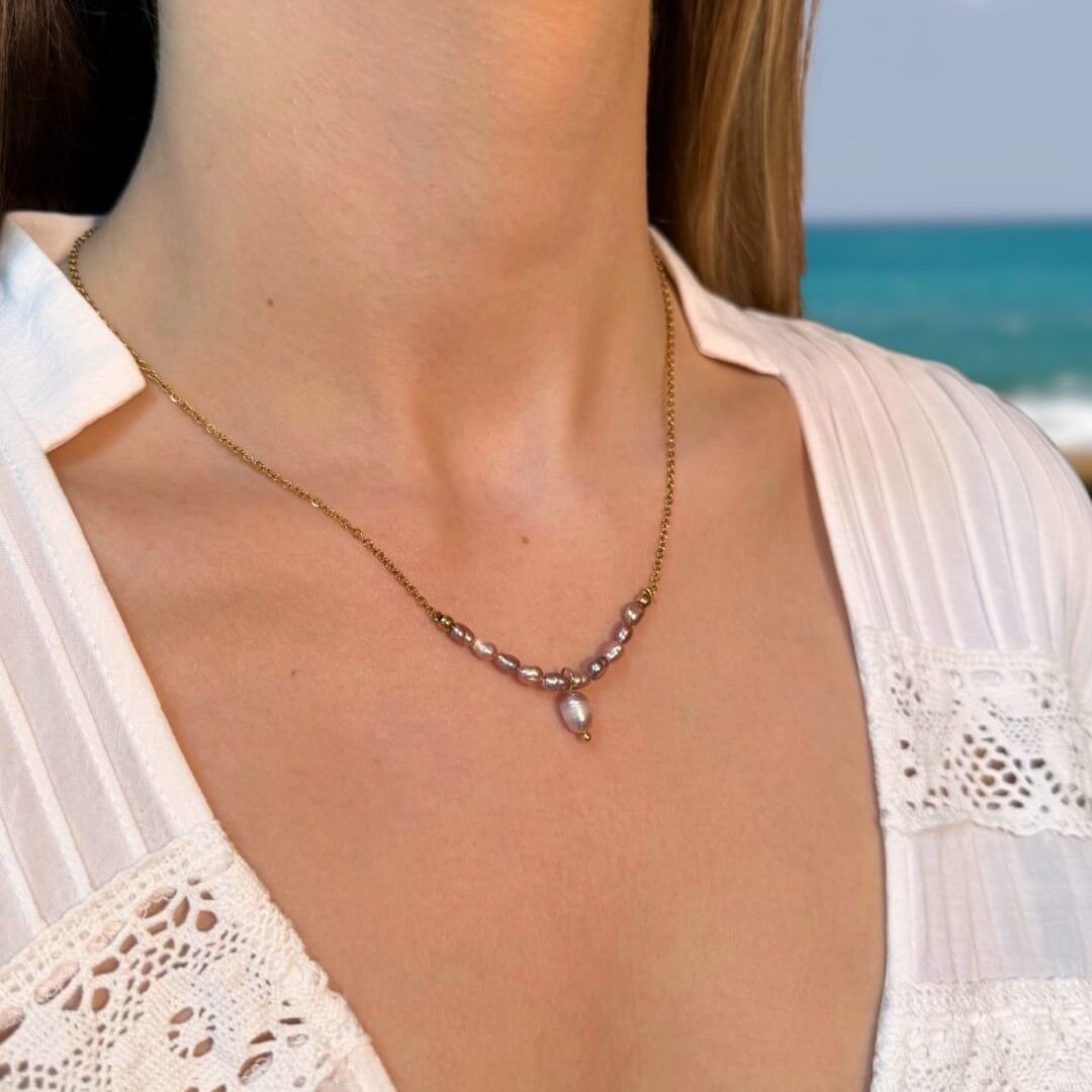Collier porté par une femme, en acier inoxydable doré avec petites perles d’eau douce et grosse perle centrale violette.