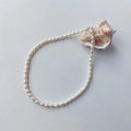 Collier ras-de-cou entièrement composé de petites perles d'eau douce blanches ovales nacrées de tailles variées, avec un fermoir argenté, disposé en cercle sur fond gris clair aux côtés d'un grand coquillage strié blanc et orangé.