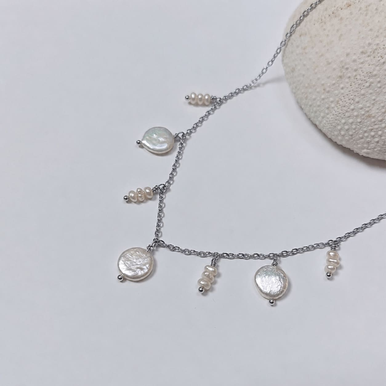 Collier en acier inoxydable argenté avec perles d’eau douce plates et petits trios de perles blanches, posé sur un corail blanc, sur fond blanc.
