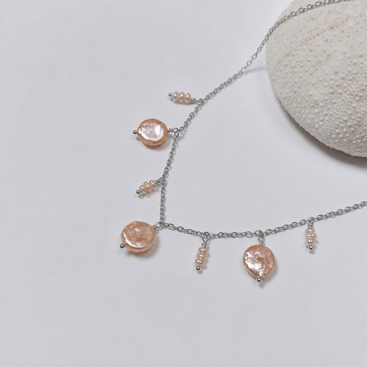 Collier en acier inoxydable argenté avec perles d’eau douce plates et petits trios de perles oranges, posé sur un corail blanc, sur fond blanc.