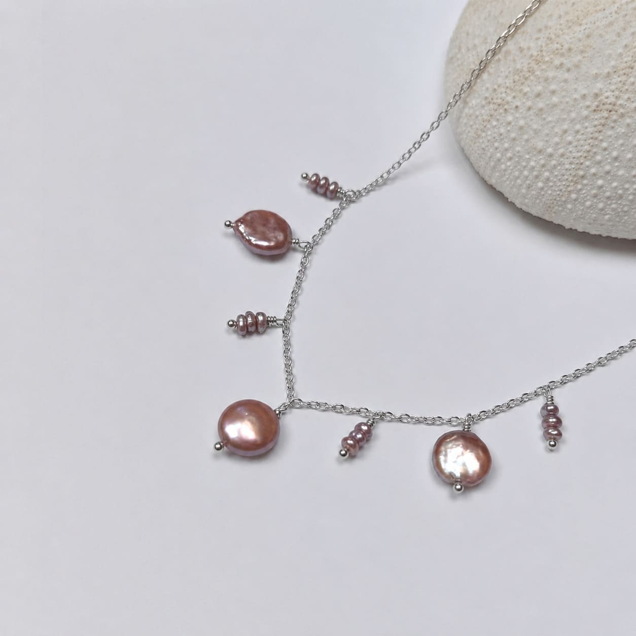 Collier en acier inoxydable argenté avec perles d’eau douce plates et petits trios de perles violettes, posé sur un corail blanc, sur fond blanc.