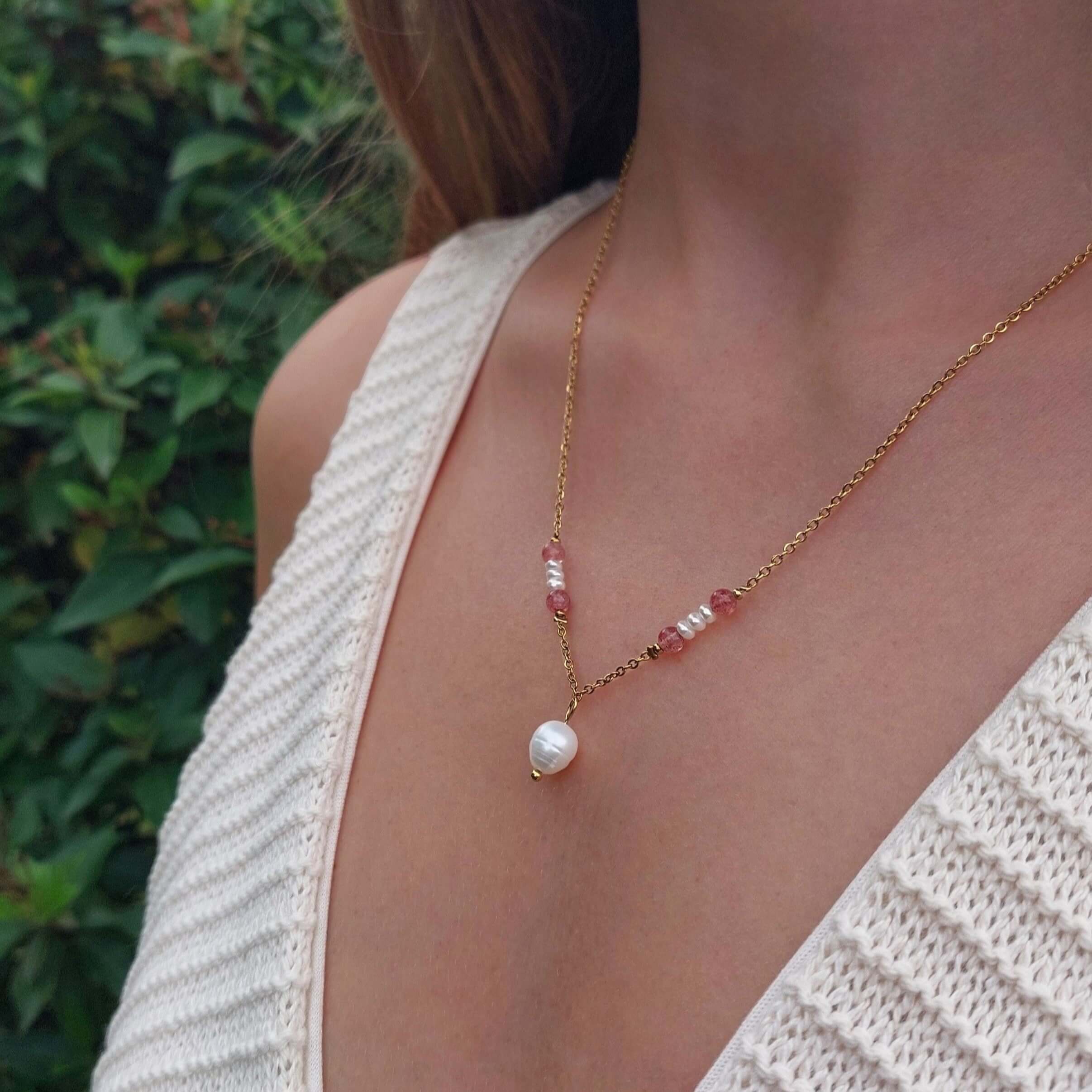 Femme portant le collier Euphyllia en quartz fraise et petites perles d'eau douce sur fine chaîne dorée en acier inoxydable, avec une grande perle d'eau douce baroque en pendentif, photographiée en extérieur avec un haut en crochet blanc et un fond de végétation verte.