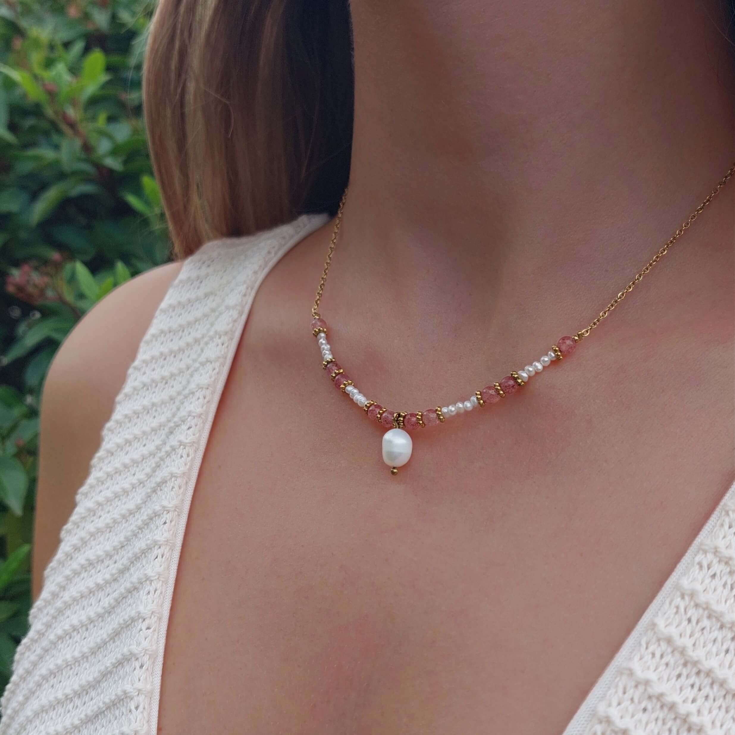 Femme portant le collier Euphyllia en quartz fraise et petites perles d'eau douce sur fine chaîne dorée en acier inoxydable, avec une grande perle d'eau douce baroque en pendentif, photographiée en extérieur avec un haut en crochet blanc et un fond de végétation verte.