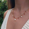Femme portant le collier Meandrina quartz fraise en acier inoxydable doré avec breloques de perles d'eau douce baroques et pierres de quartz fraise roses alternées, photographiée en extérieur avec un haut en crochet blanc et un fond de végétation verte.