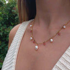 Femme portant le collier Meandrina quartz fraise en acier inoxydable doré avec breloques de perles d'eau douce baroques et pierres de quartz fraise roses alternées, photographiée en extérieur avec un haut en crochet blanc et un fond de végétation verte.