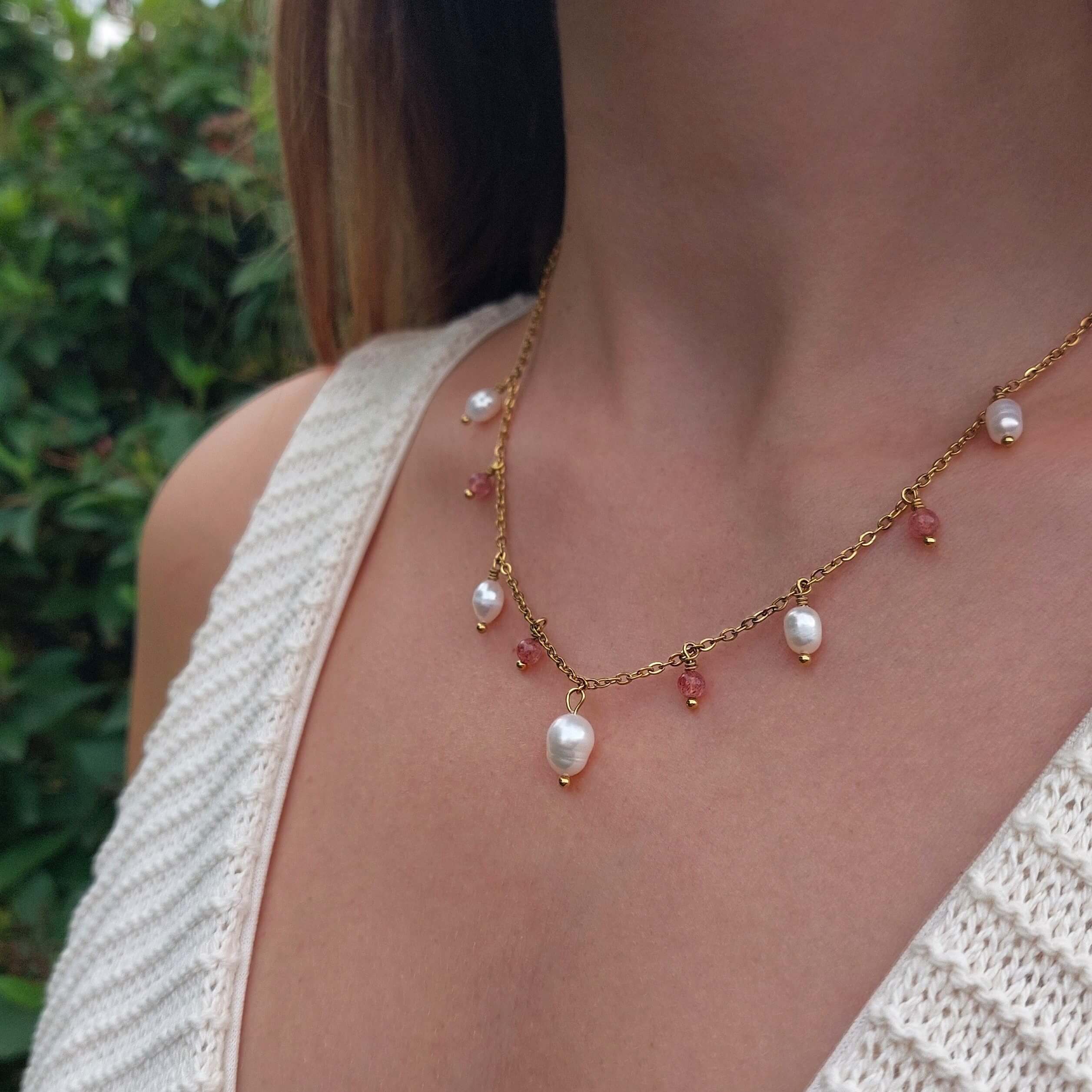 Femme portant le collier Meandrina quartz fraise en acier inoxydable doré avec breloques de perles d'eau douce baroques et pierres de quartz fraise roses alternées, photographiée en extérieur avec un haut en crochet blanc et un fond de végétation verte.