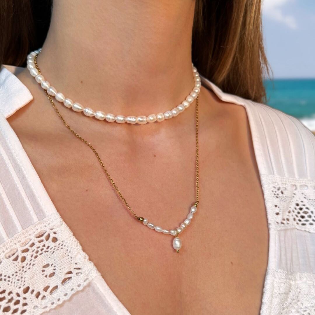 Colliers portés en accumulation par une femme ras de cou et pendant en perles d’eau douce naturelles blanches, sur un fond océan flouté. 