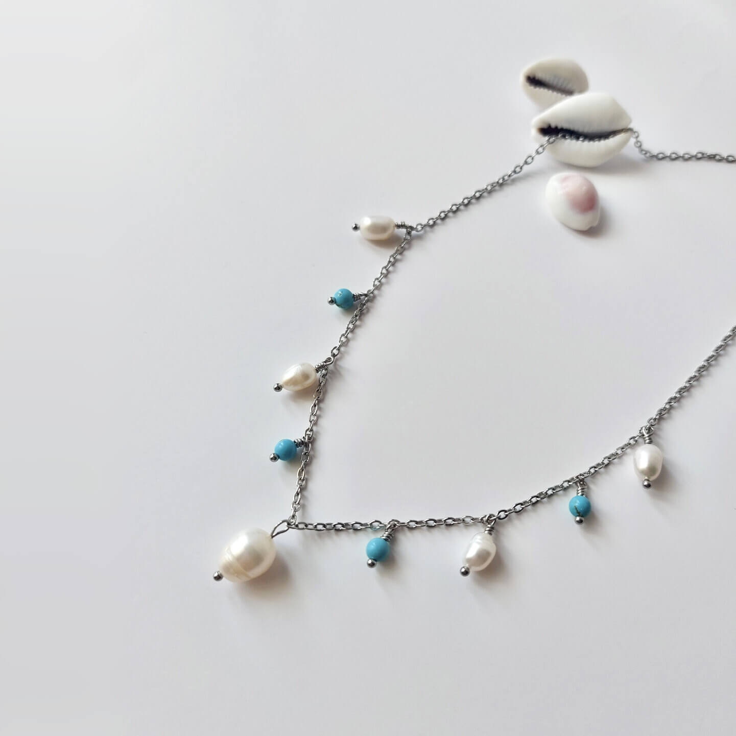 Collier Meandrina en acier inoxydable argenté, perles d'eau douce et perles de turquoise bleue posé sur fond blanc