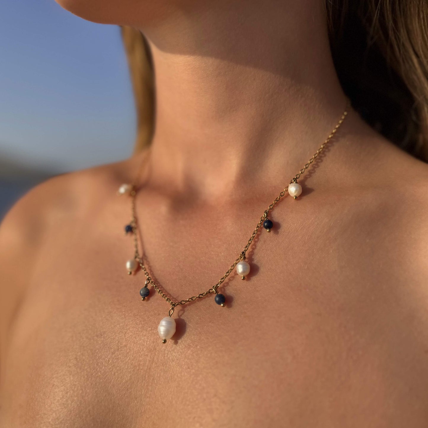 Collier sur chaine en acier inoxydable doré avec perles d'eau douce et perles de couleur porté par une femme bronzée devant la mer.