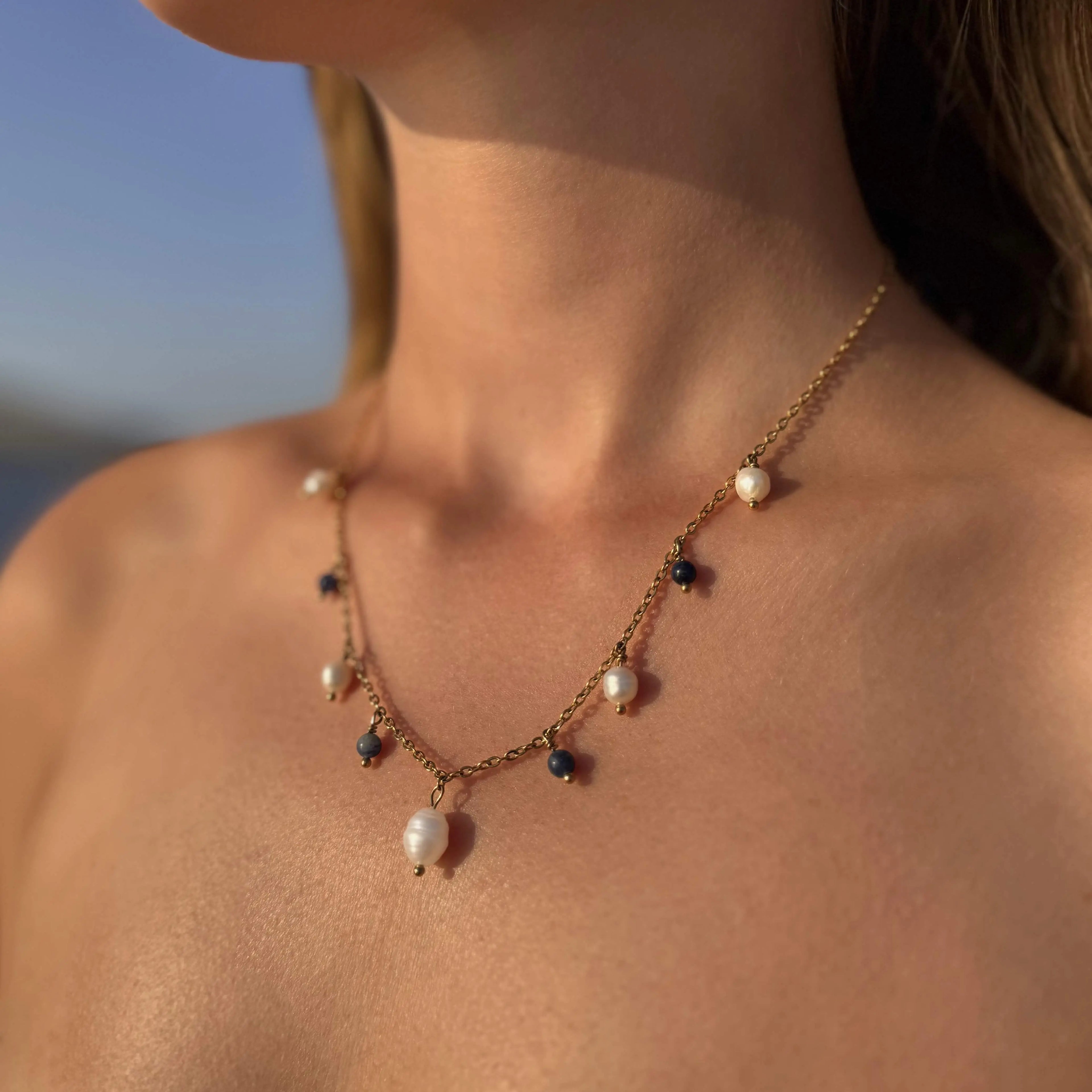 Collier sur chaine en acier inoxydable doré avec perles d'eau douce et perles de couleur porté par une femme bronzée devant la mer.