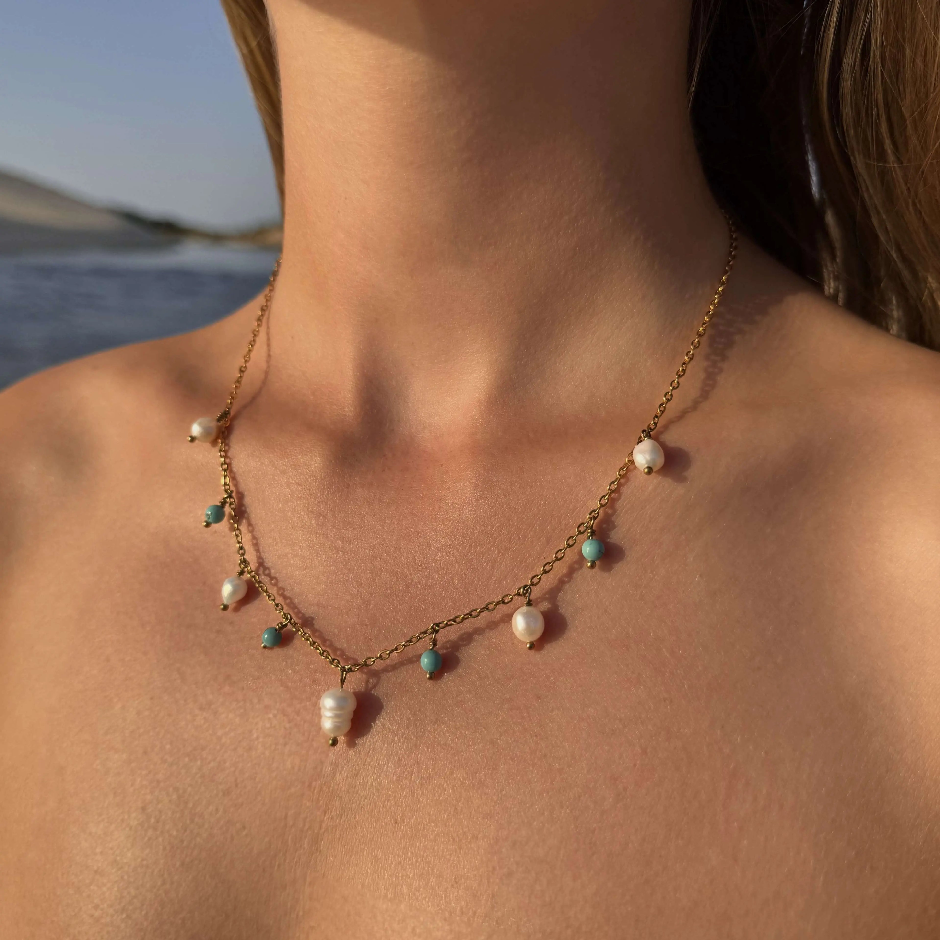 Collier sur chaine en acier inoxydable doré avec perles d'eau douce et perles de couleur porté par une femme bronzée devant la mer.