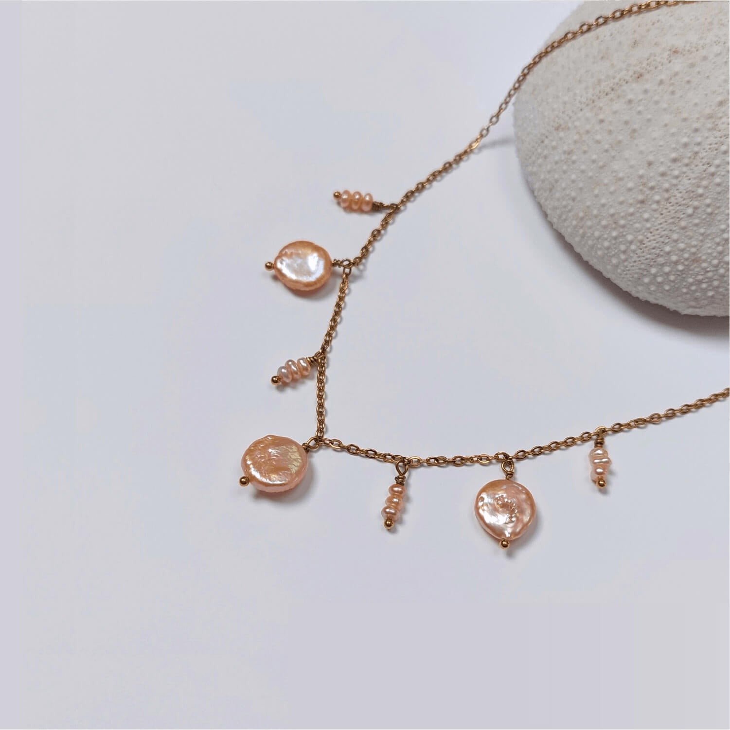 Collier en acier inoxydable doré avec perles d’eau douce plates et petits trios de perles oranges, posé sur un corail blanc, sur fond blanc.