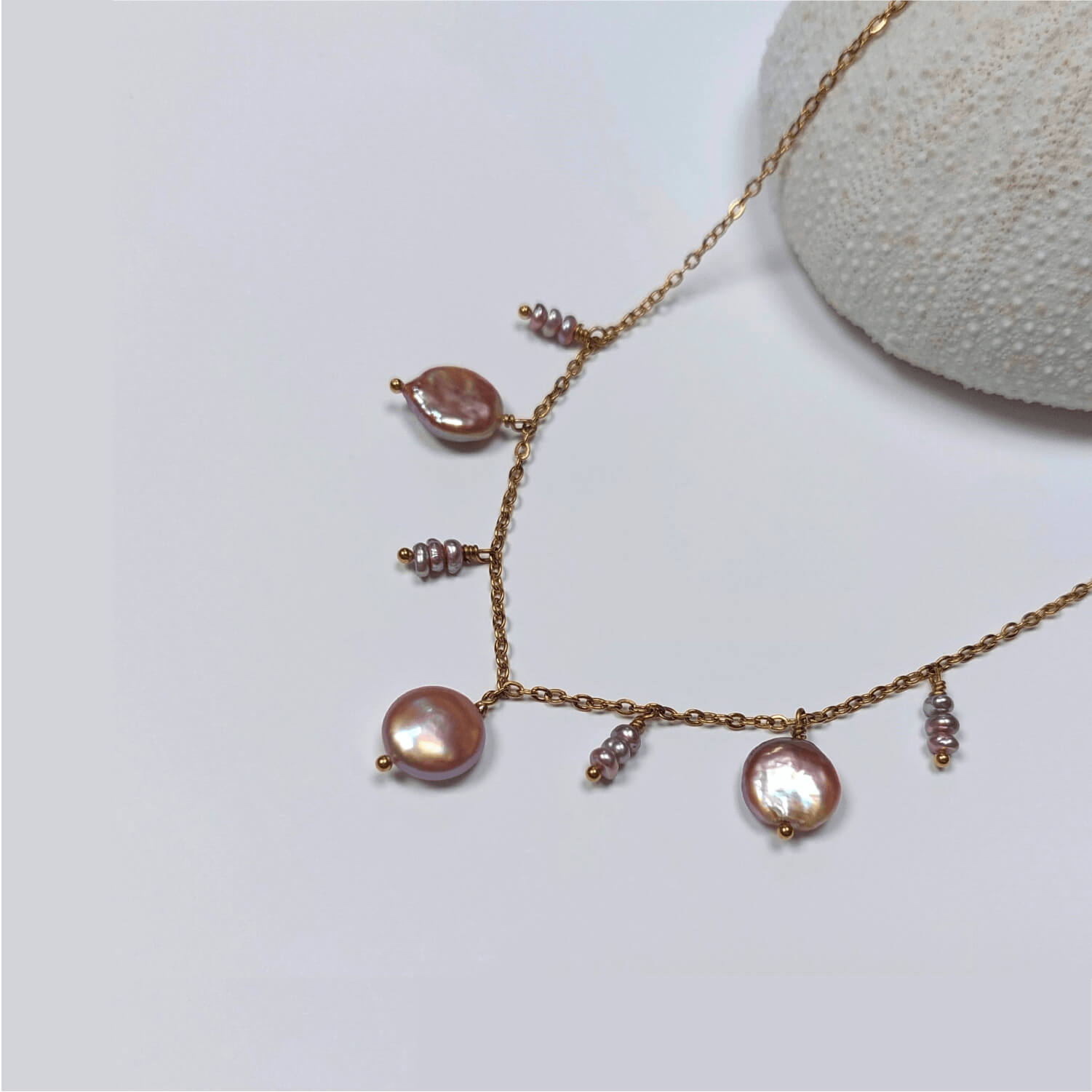 Collier en acier inoxydable doré avec perles d’eau douce plates et petits trios de perles violettes, posé sur un corail blanc, sur fond blanc.